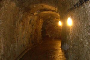 Dim underground tunnel with rough stone walls and an arched ceiling, lit by two warm yellow lamps along the right wall.