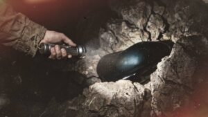 A hand holding a flashlight in a rocky cave, shining on a glossy black object nestled in a crevice.
