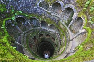 Top-down view of a moss-covered stone spiral staircase descending into a deep circular well through arched openings.