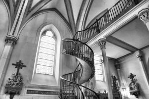 Spiral staircase inside a Gothic church with ornate railings and a tall arched window behind it.
