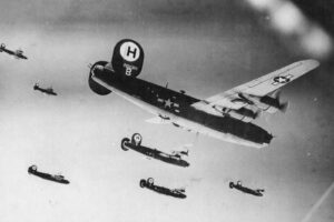 Black-and-white photo of US WWII bombers in a mid-air formation, led by a large B-17 Flying Fortress with US markings on the wings and fuselage.