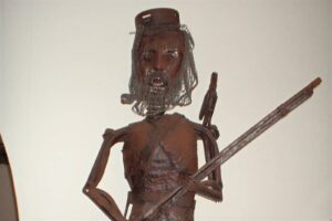 Rusty metal sculpture of a man holding a long spear, wearing a cap and wire mesh beard/hair.