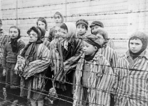 Group of children in striped clothing standing behind a barbed-wire fence, looking toward the camera.
