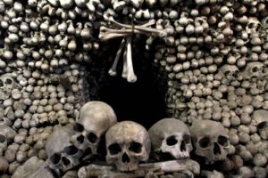 Wall of stacked human skulls and bones forming an ossuary, with a dark opening in the center and a few skulls resting at the front.