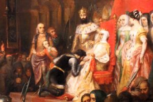 Stage scene from a royal drama: king in crown stands beside a seated queen on a red throne, surrounded by courtiers in ornate costumes; audience visible in foreground.