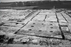 Aerial view of a large grid-like concrete foundation with rectangular slabs and evenly spaced holes on a barren coastal construction site near water.