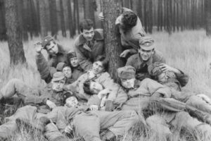A group of soldiers in uniform relaxing on the grassy ground around a tree in a pine forest, some with cigarettes.