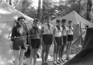 Seven scouts in uniform stand in a line at a forest campsite with canvas tents behind them.