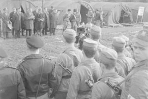 Group of soldiers in uniform with rifles stands in a line at a muddy camp, while civilians with documents and a photographer observe from behind them near canvas tents.