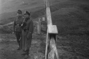 Two soldiers in historic military uniforms stand beside a tall wooden cross on a hillside grave marker in a remote landscape.