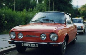 Front view of a red vintage Škoda car with four round headlights and a European license plate reading OU 43-48 on a suburban street.
