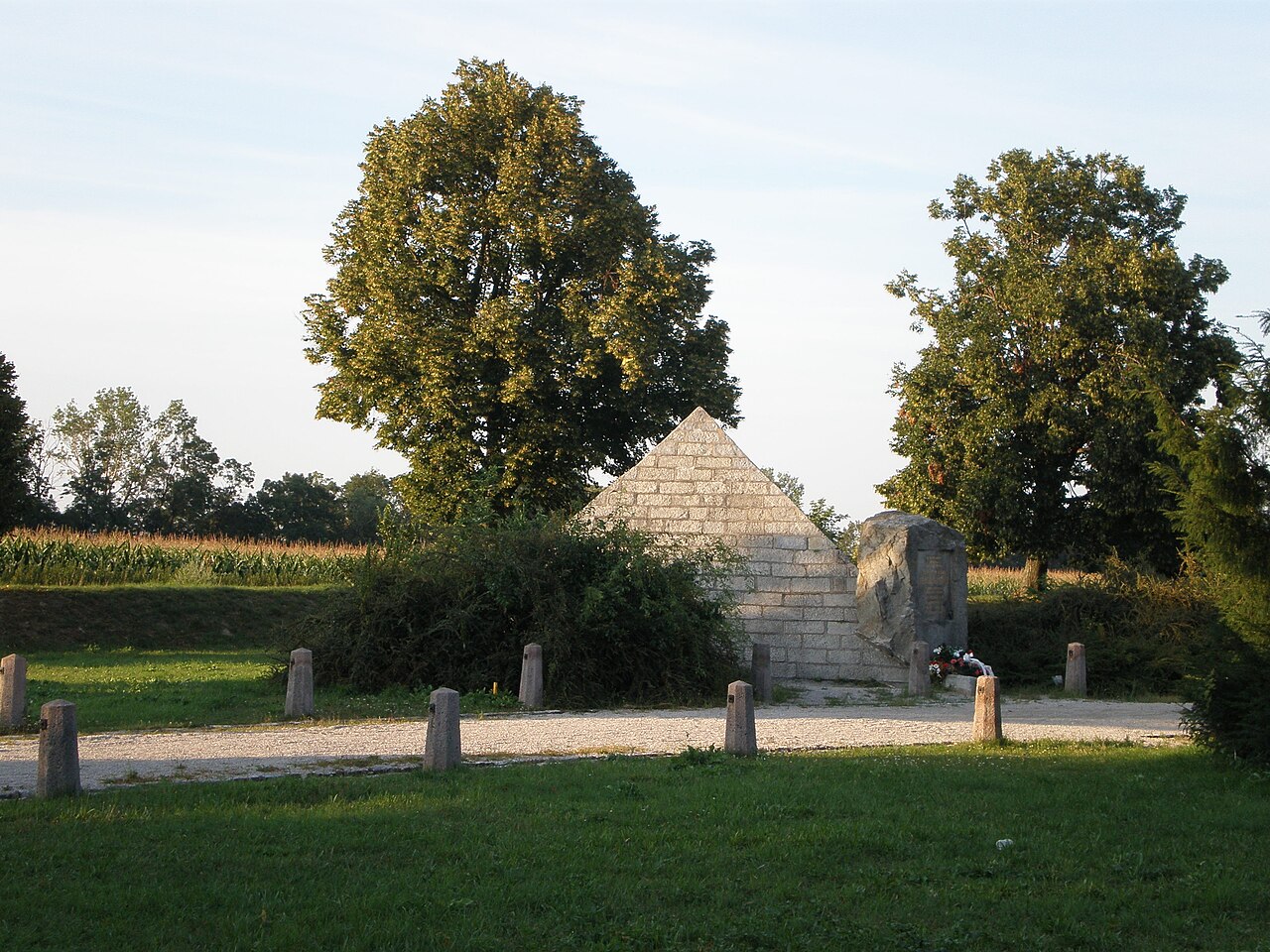 Stone pyramid memorial in a sunlit park, with a gravel path and trees in the background.