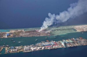 Aerial view of a coastal harbor with industrial buildings and a large plume of white smoke rising from the land area.