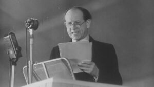 Man in a suit and glasses delivering a speech at a lectern, reading from held notes with a vintage microphone nearby.