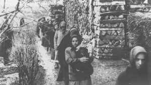 Line of women in headscarves walking along a path beside stacked firewood, one woman at the front holds a baby. captures a refugee or displacement scene