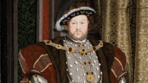 Portrait of a Tudor king in opulent robes, heavy embroidery, and gold jewelry, posed beside patterned drapes