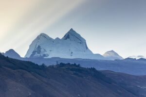 Snowy mountain peak rising above rolling hills with a pale blue sky and distant ridges to the right.
