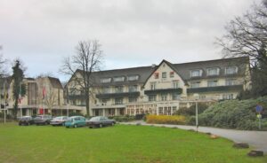 Hotel De Bilderberg exterior with flags, a lawn, and parked cars in front of a multi-story building under cloudy sky