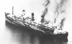 A large ship on fire with flames and smoke rising from the deck at sea (black and white image).