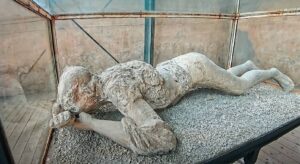 Life-size stone sculpture of a figure lying on its side inside a glass display case, with a rough weathered texture on a gravel base.