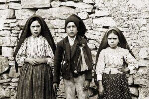 Three children in traditional clothing stand against a rough stone wall; two girls wear shawls, the center child wears a coat and hat.