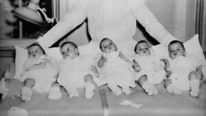 Five newborn babies lie on a hospital bed in a row, dressed in white, with a caregiver seated behind them.