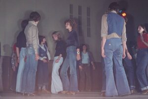 A group of young people in a gym or studio, wearing 70s-style denim jeans and casual tops, facing each other as if rehearsing or chatting.