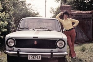 Woman in yellow blouse and striped pants poses beside a light vintage car, leaning on the car's fender with one arm raised.
