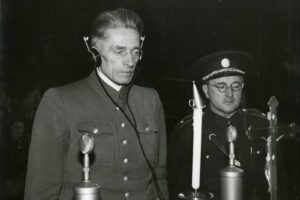 Two men in vintage military uniforms speak into old-fashioned microphones on a stage. One wears headphones and looks down; the other, in a peaked hat, stands beside him.