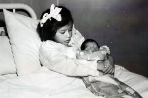 Young girl with a large white bow in her hair sits in a hospital bed, cradling a newborn baby wrapped in a blanket.
