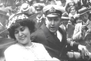 Bride in a white dress with a floral crown smiles beside a man in a naval captain’s cap, amid a cheering crowd in vintage clothing and hats at a parade or public celebration.