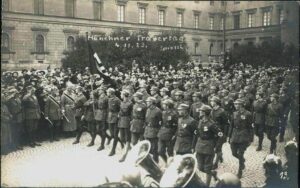 Hundreds of soldiers march in formation along a cobblestone street while crowds watch; a banner overhead reads 'Monheimer Trauertage'.