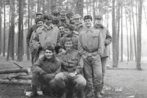 Group of young soldiers in uniform posing together in a forest clearing, several smiling, with logs on the ground behind them.