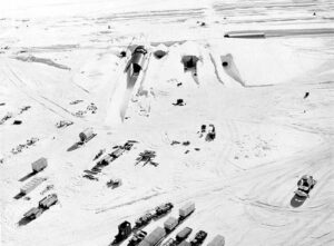 Snow-covered industrial field with rows of containers and equipment around a large snow mound and tunnels.