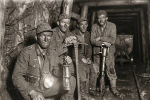 Five miners in a coal mine, wearing dirty uniforms and hats, holding lanterns and mining tools, kneeling and leaning against a rocky wall.