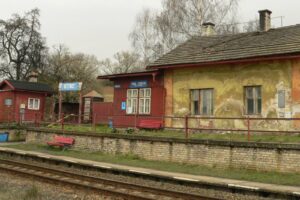 Old rural train station with peeling yellow walls, red wooden structures, a platform, and parallel tracks.