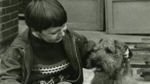 Young child in a jacket pats a fluffy, scruffy dog outdoors beside a brick wall.
