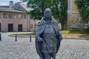 Statue of a medieval king in a cloak standing in a cobblestone town square with pastel buildings and green trees behind.