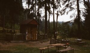 Wooden shrine in a forest clearing, with a cross on a tall post and a fence decorated with white ribbons.
