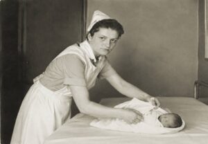 Nurse in a white uniform and cap tends to a newborn wrapped in a blanket on a hospital bed.