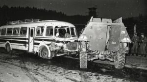 Night scene of a damaged white bus crashed into a large armored military vehicle; soldiers stand nearby in the background.