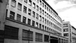 Black-and-white photo of a multi-story brick building with many rectangular windows along a street; a streetlamp stands in the foreground.