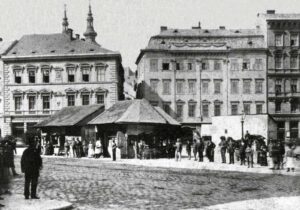 Historic European town square with a market canopy and people queuing, set against ornate multi-story buildings.