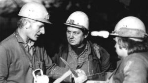 Three miners wearing hard hats discuss while one holds a tool in a dim underground setting (black and white).