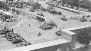 Aerial view of a city intersection crowded with tanks and military vehicles, with soldiers and civilians observing from the sidewalks.