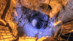 Deep vertical mine shaft surrounded by rough rock, with metal railing along the edge and a blue-tinged glow down the hole