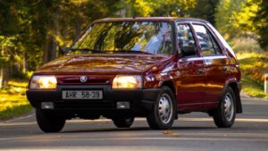 Burgundy compact car driving on a sunlit, tree-lined road with autumn leaves nearby.