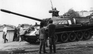 Black-and-white photo of a tank with a long cannon on display, civilians and soldiers standing nearby on a street.