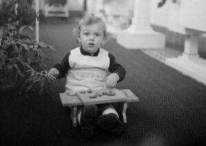 Toddler wearing a light apron sits on a small wooden ride-on toy with a tray of toy fruits outdoors, gazing ahead with wide eyes.