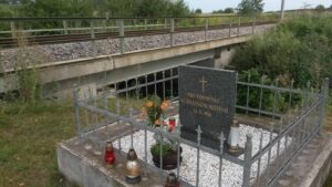 Small grave enclosed by a gray metal fence, with a polished stone headstone and flowers beside a railway embankment.
